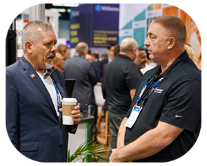 Two men engaged in conversation at a bustling trade show, surrounded by various booths and attendees