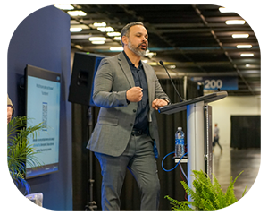 A man in a suit delivers a speech at a convention, engaging the audience with his presentation