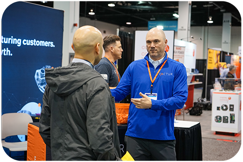 Two men engaged in conversation at a bustling trade show, surrounded by various booths and attendees