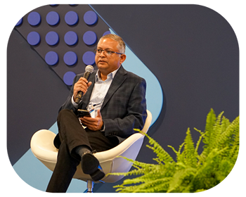 A man seated in a chair, speaking into a microphone during a presentation or interview
